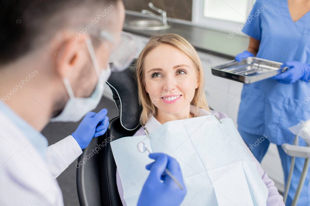 Happy young woman looking at her dentist with toothy smile while sitting in front of him before oral examination