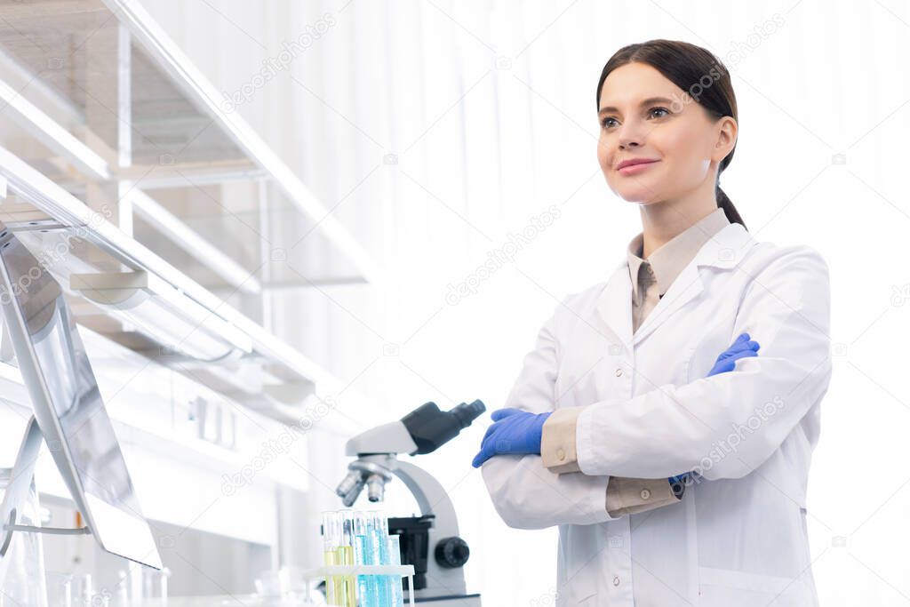 Confident Caucasian female doctor wearing white lab coat and gloves standing with arms crossed in modrn laboratory