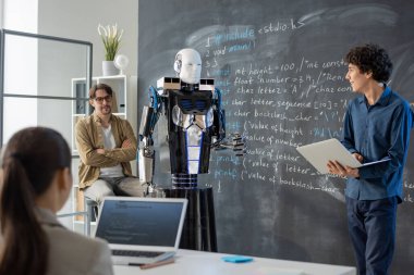 Sınıfta dizüstü bilgisayarı olan çağdaş genç bir öğrenci karatahta duruyor ve bilgisayar robotu sunumu yapıyor.