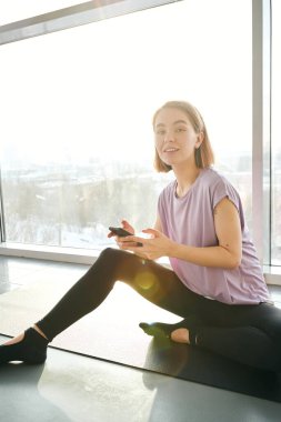 Akıllı telefonlu genç bir bayan kameranın önünde büyük bir pencerede oturuyor ve antrenman sırasında sana bakıyor.