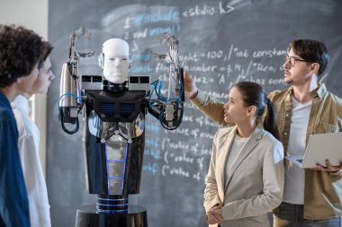 Bir grup öğrenci ellerini kaldırmış bilgisayar kontrol robotuna bakarken öğretmenleri derste yeteneklerini gösteriyor.