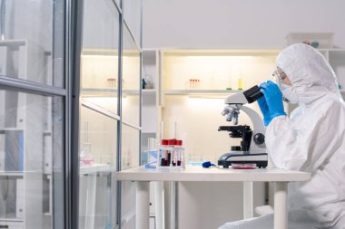 Side view of medical scientist in protective mask using microscope while studying dangerous coronavirus in laboratory