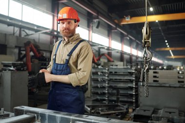 Fabrika atölyesinde, iş kıyafetleri ve başlık konusunda ciddi bir mühendis kameranın önünde büyük bir demir parçasının yanında duruyor.