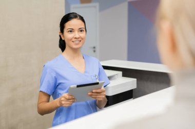 Dijital tabletli genç bir asistan kliniklere yeni hasta kaydedilirken resepsiyonda bekliyor.