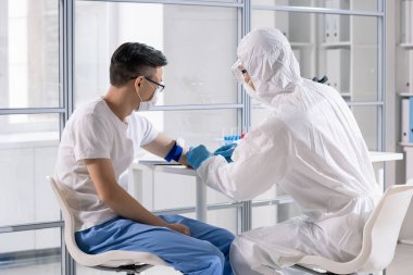 Medical specialist in biohazard suit taking blood of deadly infected Asian patient in control area