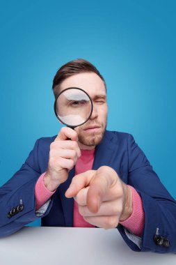 Portrait of serious smart young man pointing at camera and looking through magnifying glass while investigating you clipart