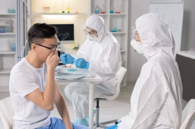 Virology specialist in biohazard suit and respiratory mask talking to coronavirus patient in isolated research room