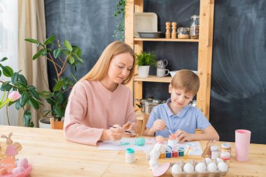 Genç anne ahşap masada oturuyor ve Paskalya partisi için yumurta boyamasına yardım ediyor.