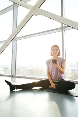 Bir bacağı esnemiş genç bir sporcu geniş bir pencerede oturuyor ve akıllı telefondan yoga videosu izliyor.