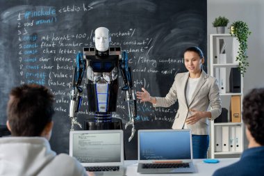 Akıllı genç kız öğrenci karatahtanın yanında dururken sınıf arkadaşlarına yeni bir otomasyon robotu modeli gösteriyor.