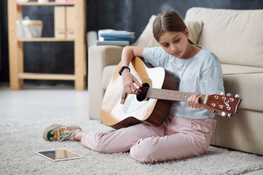 Kendi kendine öğrenen genç kız halının üzerinde oturuyor ve gitar çalarken eğitici videolar kullanıyor.