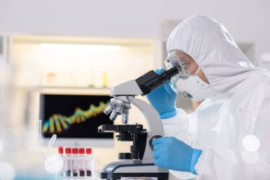 Side view of researcher in protective suit and mask using microscope while studying virus in laboratory