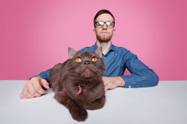 Ciddi bir kedi sahibinin portresi, pembe arka planda kahverengi evcil hayvanla masada oturuyor.