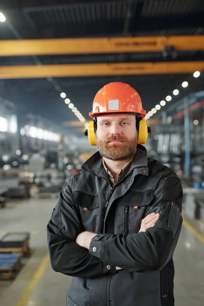 工厂里快乐的年轻大胡子产业工人 头戴防护耳机 头戴头盔 站在摄像机前 — 图库照片