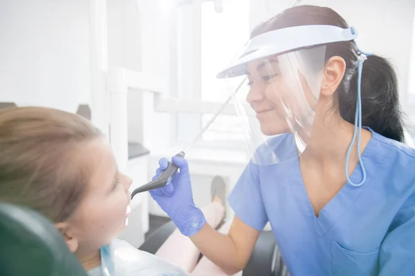 戴防护面具 戴手套 身穿蓝色制服的年轻女牙医将在口试后修补小女孩的牙齿 — 图库照片