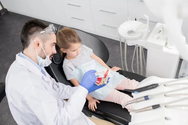 年轻的牙医戴着手套 满嘴脏话 一边指着假牙一边向小女孩解释他要做什么 — 图库照片