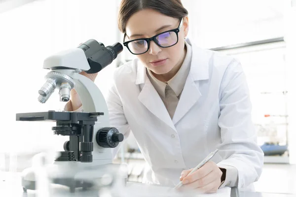 Jovem Trabalhadora Médica Fazendo Anotações Após Examinar Espécime Usando Microscópio — Fotografia de Stock