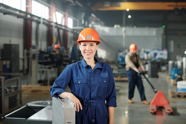 工場の倉庫に立っている間あなたを見てハードハットとブルーのカバーで幸せな若い笑顔の女性エンジニア — ストック写真