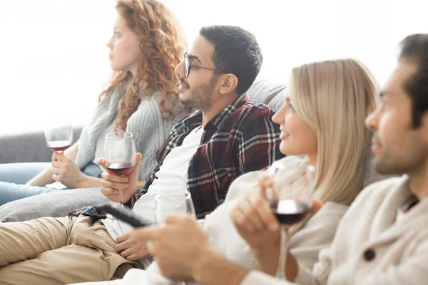 Group of young multi-ethnic friends with wineglasses sitting on sofa and watching tv together