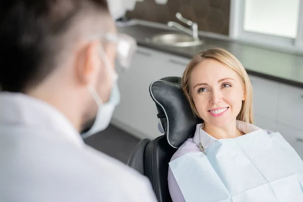 幸せな若いですブロンド女性患者見ます彼女の歯医者とともに健康的な笑顔とともに座っていますアームチェア前に試験 — ストック写真