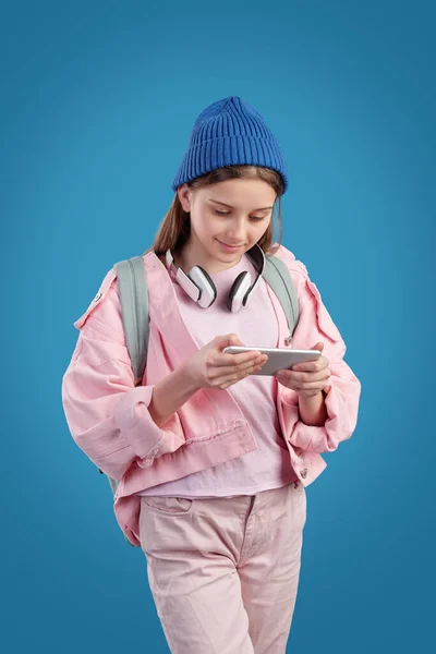 Curious hipster teenage girl in pink jacket wearing wireless headphones on neck watching video on phone