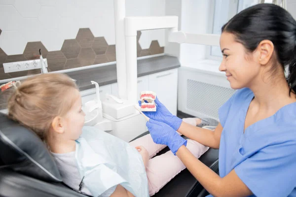 穿着蓝色制服和手套的年轻的黑发助手向小女孩展示假牙 并向她解释如何正确地刷牙 — 图库照片
