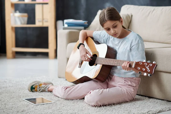 独学の十代の女の子はカーペットの上に座って ギターを演奏しながらタブレット上で指導ビデオを使用して — ストック写真
