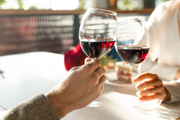 Horizontal close up shot of unrecognizable man and woman sitting in restaurant clinking glasses with red wine