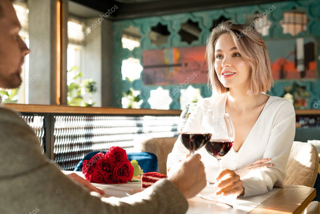 Horizontal over-the-shoulder shot of young adult man and woman in love sitting in restaurant clinking glasses with red wine