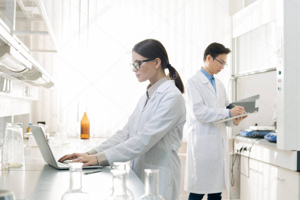 Horizontal shot of young man and woman wearing white coats standing in modern laboratory working with documents