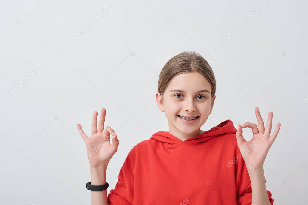 Portrait of teenage girl with dental braces wearing red hoodie showing ok sign against white background