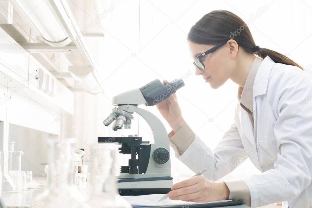 Horizontal side view shot of young female medical scientist examining specimen using microscope in modern laboratory