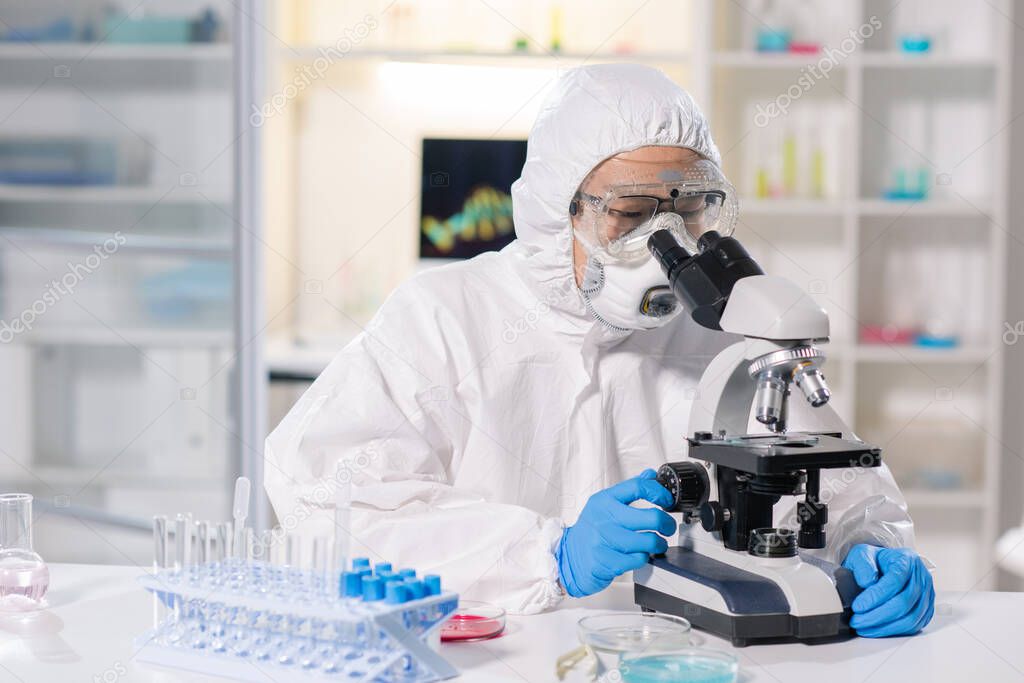 Asian medical scientist in respirator and safety goggles using microscope to research infected blood sample in laboratory