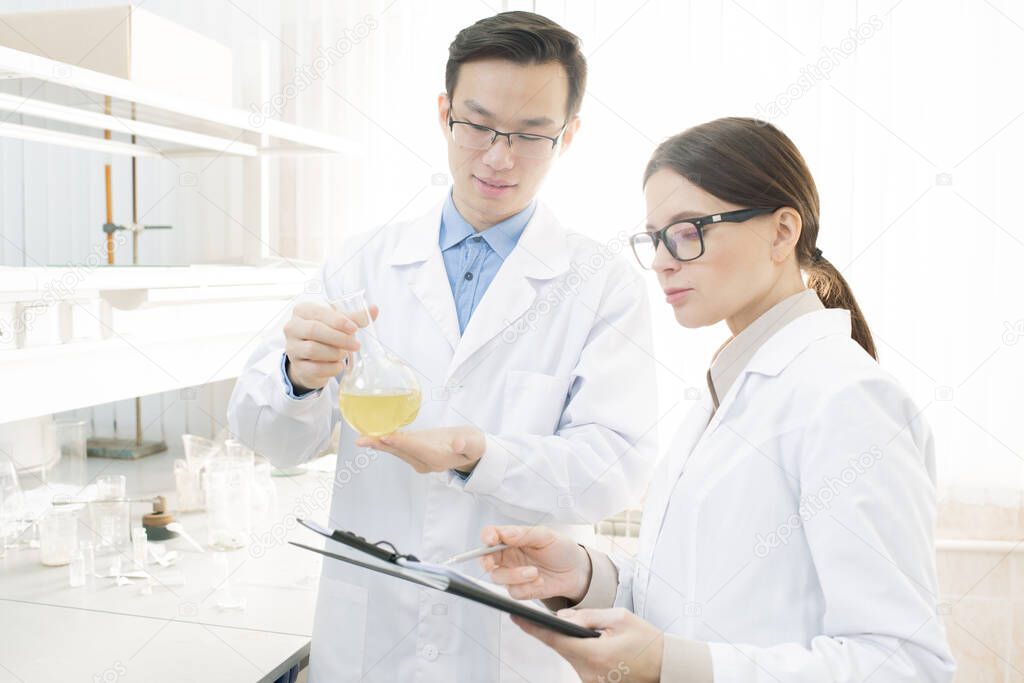 Two young medical scientists in white coats conducting research in modern laboratory, horizontal shot