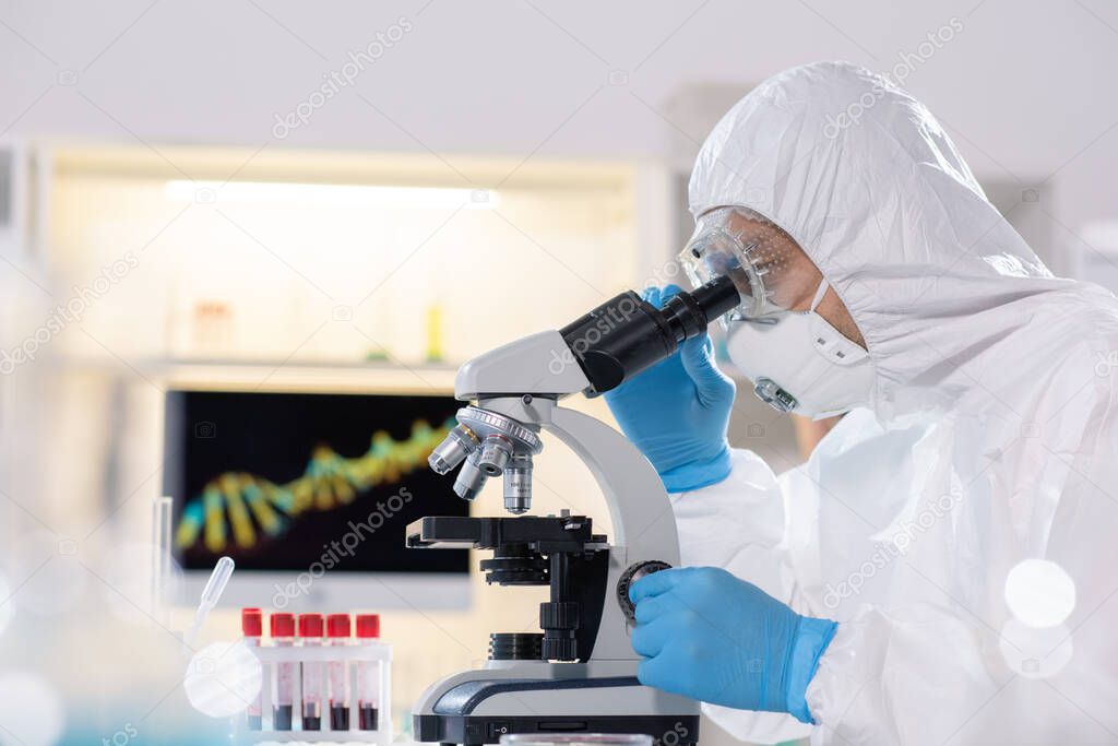 Side view of researcher in protective suit and mask using microscope while studying virus in laboratory