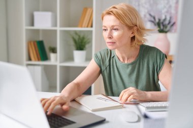 Yetişkin, sarışın, göz altı bantlı, evde tek başına çalışırken dizüstü bilgisayara bakan bir kadın.