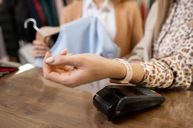 Bileğinde akıllı bilekliği olan genç bir tüketici kollarını tezgahın üzerinde tutuyor ve butikte kıyafetlerin parasını ödüyor.