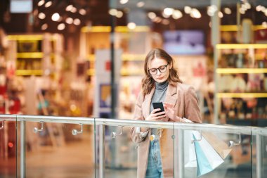 Gözlüklü, ciddi bir genç kadın alışveriş merkezinde parmaklıklar arkasında dikilip akıllı telefonları kontrol ediyor.