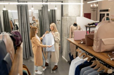Butikte ya da alışveriş merkezinde yeni kıyafetler seçerken olgun bir kadına sweatshirt gösteren genç bir kadın.