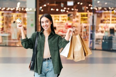 Alışveriş merkezinde alışveriş yaparken kredi kartı kullanan, kağıt torbalı heyecanlı genç bir kadının portresi.