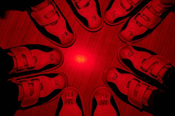 Feet of five friends in bowling shoes standing close to one another and making circle on the floor backlit with red light