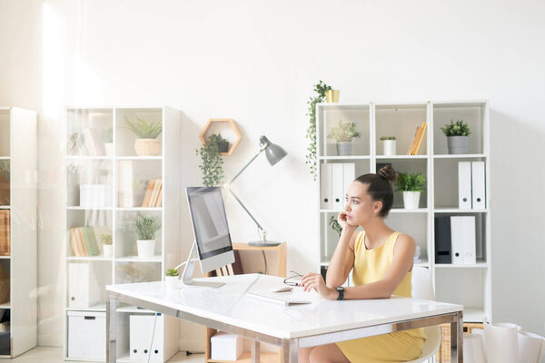 Young pensive businesswoman in elegant yellow dress looking at computer screen while organizing work or taking short break