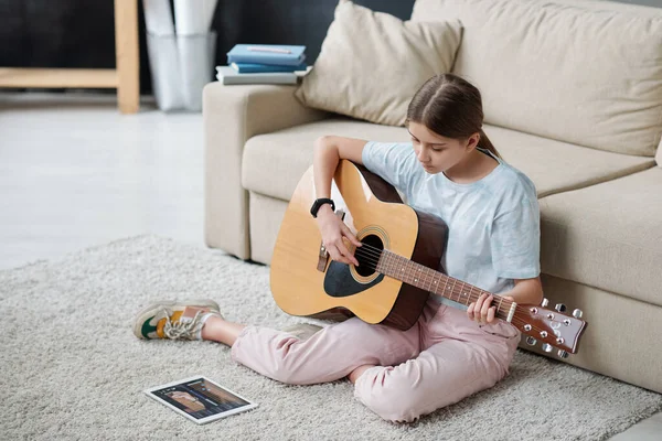 Pretty girl sitting on the floor by couch and learning to play guitar while looking at touchpad screen during online lesson