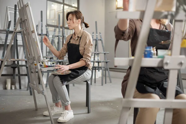 Young students of professional painting course with color palettes and paintbrushes sitting in front of easels and working
