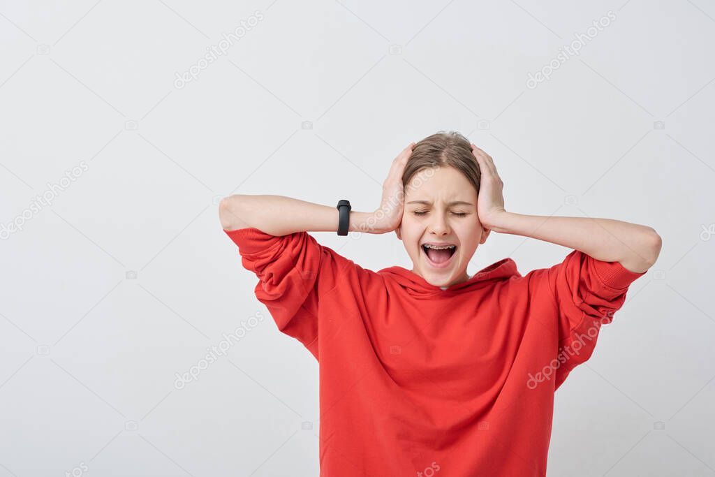 Shocked or hysteric teenage girl in red hoodie covering her ears while touching head and screaming against white background