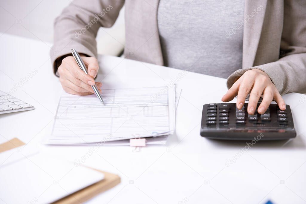 Hands of young elegant accountant with pen over financial document pressing buttons of calculator while working by desk