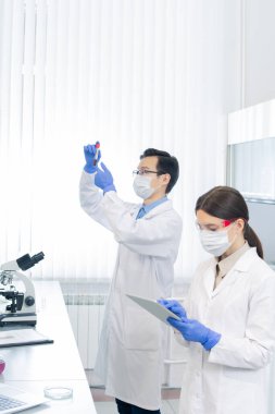 Meslektaşı laboratuarda yeni bir aşı ya da kimyasal madde içeren matara ararken koruyucu iş kıyafetleri içinde genç bir kadın tablette kayıyor.