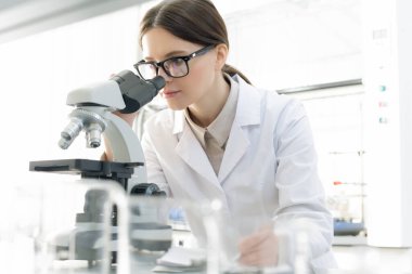 Bilim laboratuarında kovid19 üzerinde çalışırken mikroskop altında mikroskoba bakan gözlüklü genç bayan araştırmacı.