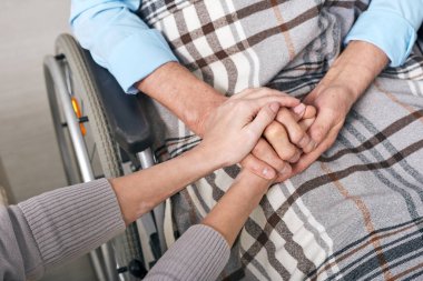 Close-up of unrecognizable woman holding hands of senior man sitting under blanket in wheelchair clipart