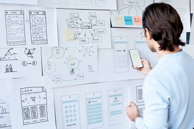 Ofiste arayüz tasarımı üzerinde çalışırken akıllı telefondaki yeni uygulamayı test eden kahverengi saçlı adamın arka görünümü